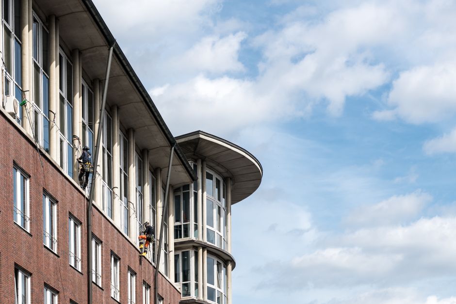 Two professional window cleaners from Cleaners Highbury are suspended on a suspended platform, performing surface cleaning on the exterior glass windows of a tall high-rise building with a brick facad