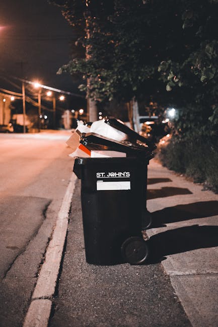 A sanitation worker wearing a high-visibility yellow and red vest is cleaning the rear compartment of a large red waste collection truck parked on the side of a street. The truck, marked with official