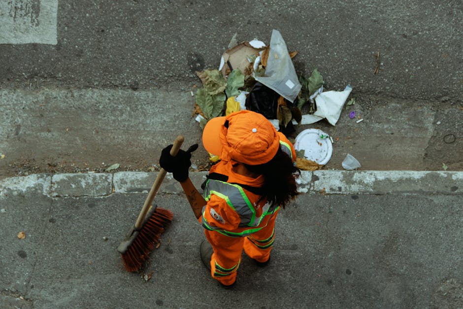 A person wearing an orange protective jumpsuit and a black respirator mask stands outdoors against a concrete wall, holding two large green waste bags filled with waste. The individual is dressed in w